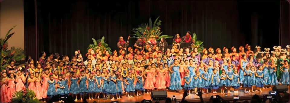 A panoramic group photograph of the hālau’s keiki across the stage in blue, pink and green hula attire, with musicians behind them.