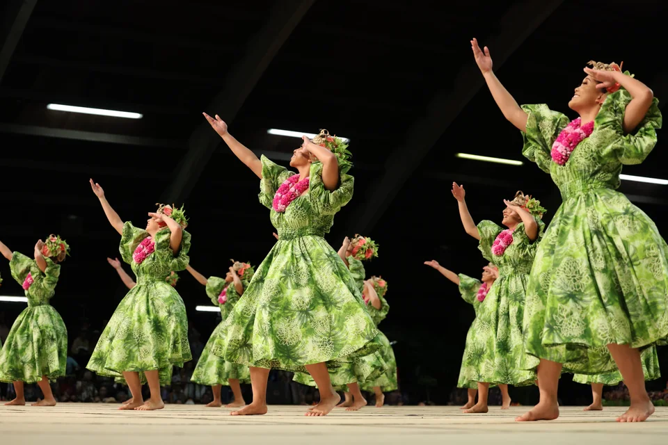 Hālau dancers in bright green dresses and pink lei, arms extended in dance.