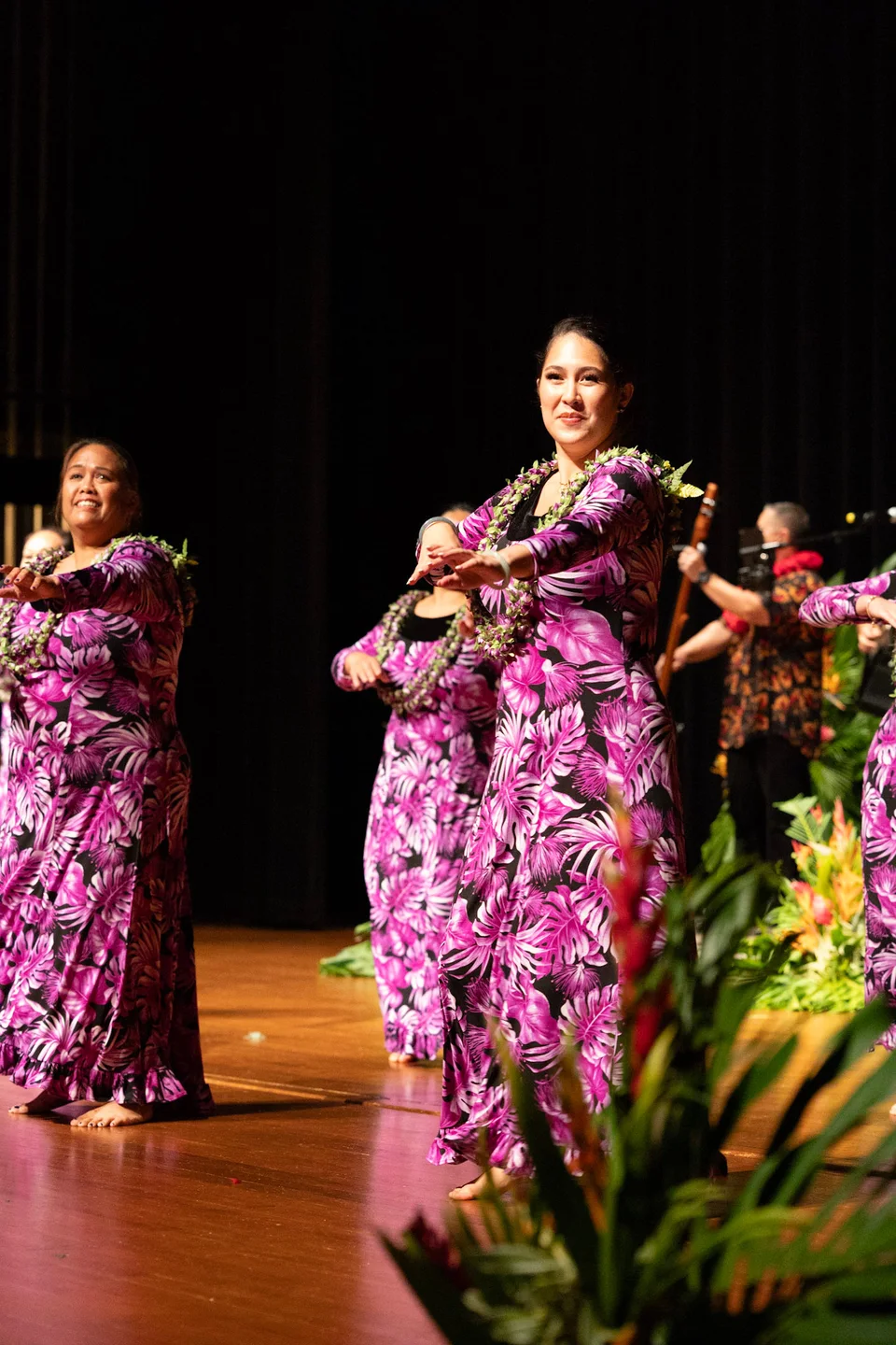 Two hula dancers performing on stage in purple floral dresses.