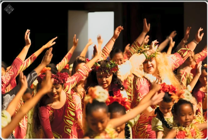 Kumu Shelsea Lilia Ai and Kumu Olana Ai sharing a hula performance with dancers of different generations, arms raised together.